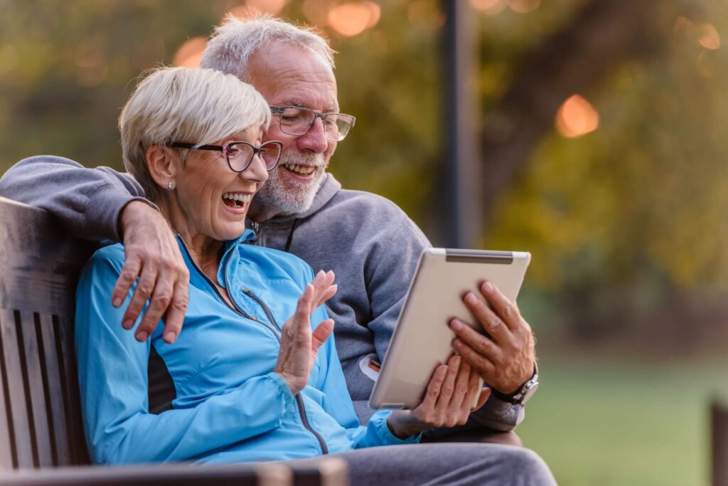 Man & woman sitting outdoors on bench looking on their tablet smiling