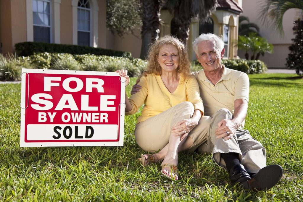 Man & woman sitting on their lawn in front of a for sale sign by owner