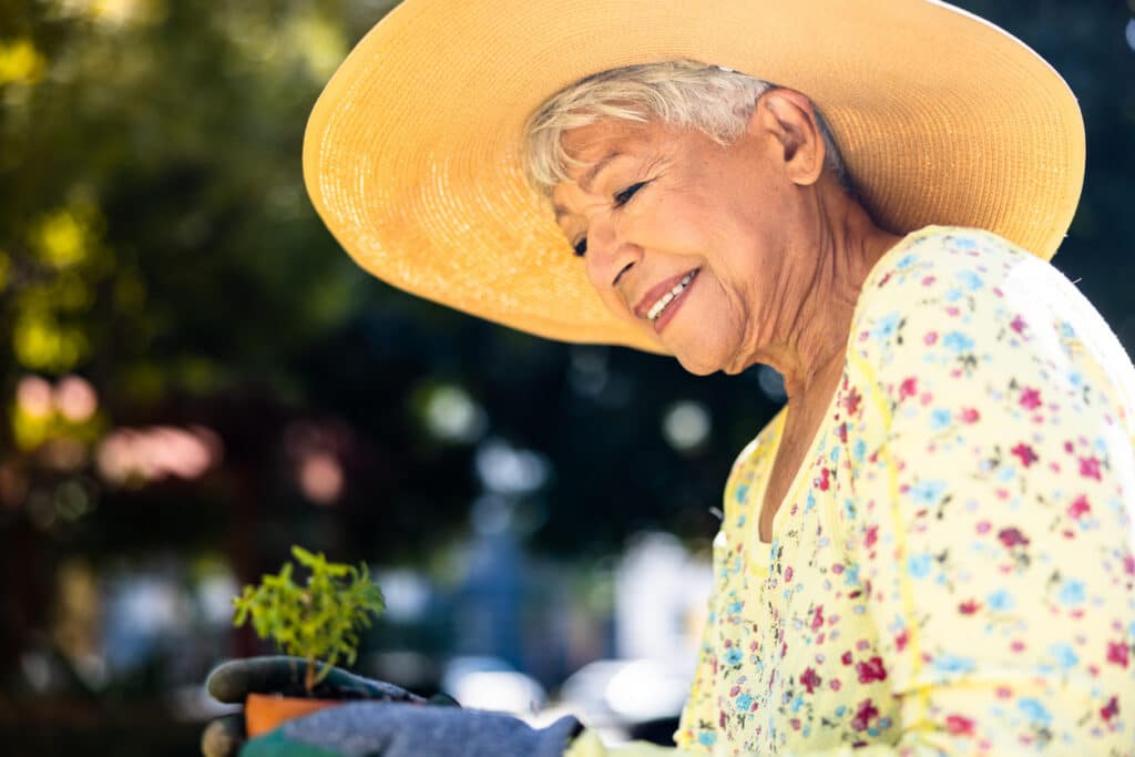 A senior woman is outside wearing a hat and gloves, admiring a plant