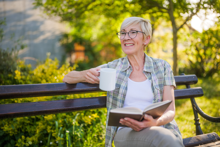 A senior woman is sitting outside on a park bench, reading and drinking coffee