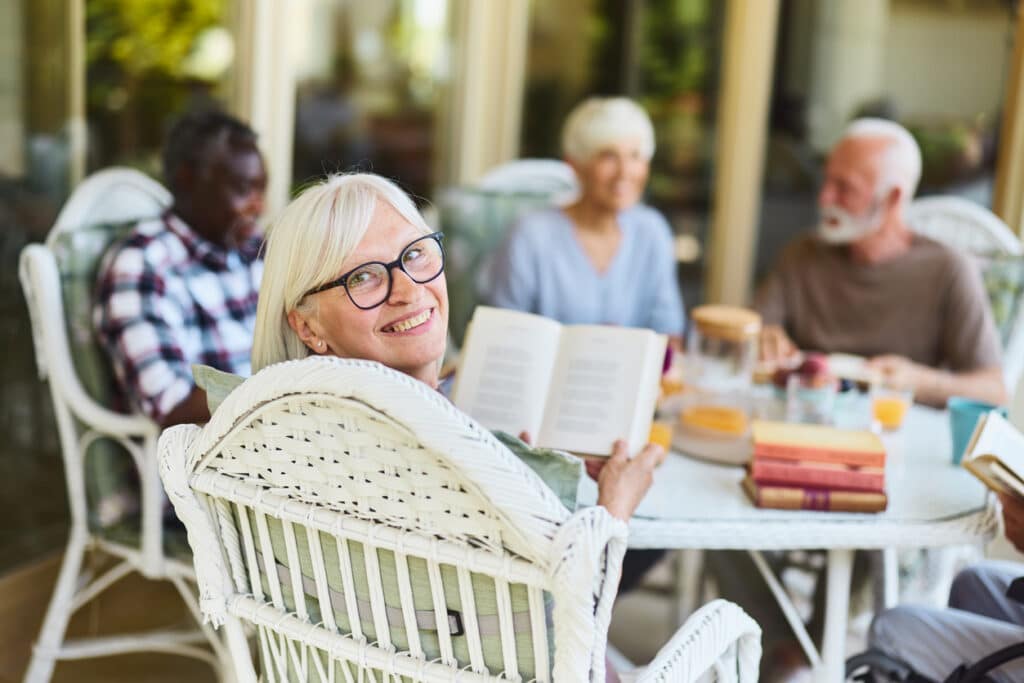 Happy mature woman reading a book while enjoying with her friends during social gathering on a balcony of a nursing home and looking at camera.